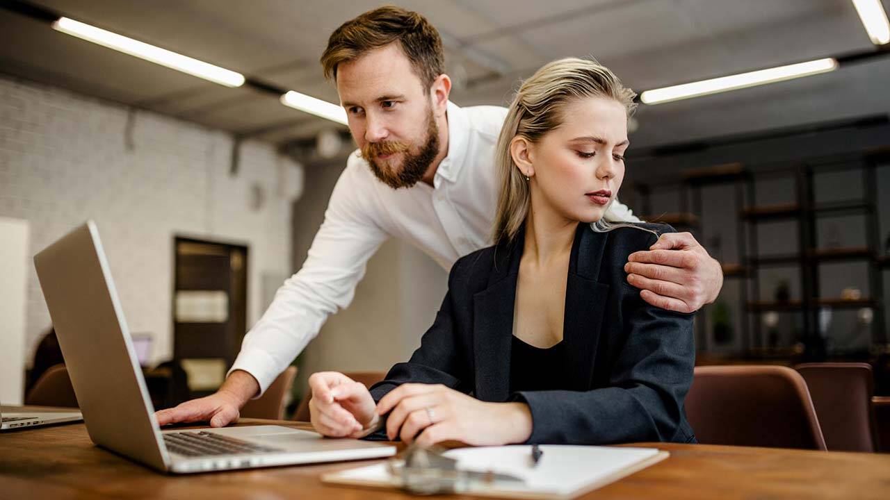 A man stands beside a seated woman in an office setting. The woman appears uncomfortable as the man places his hand on her shoulder. A laptop and notepad are on the desk in front of them.