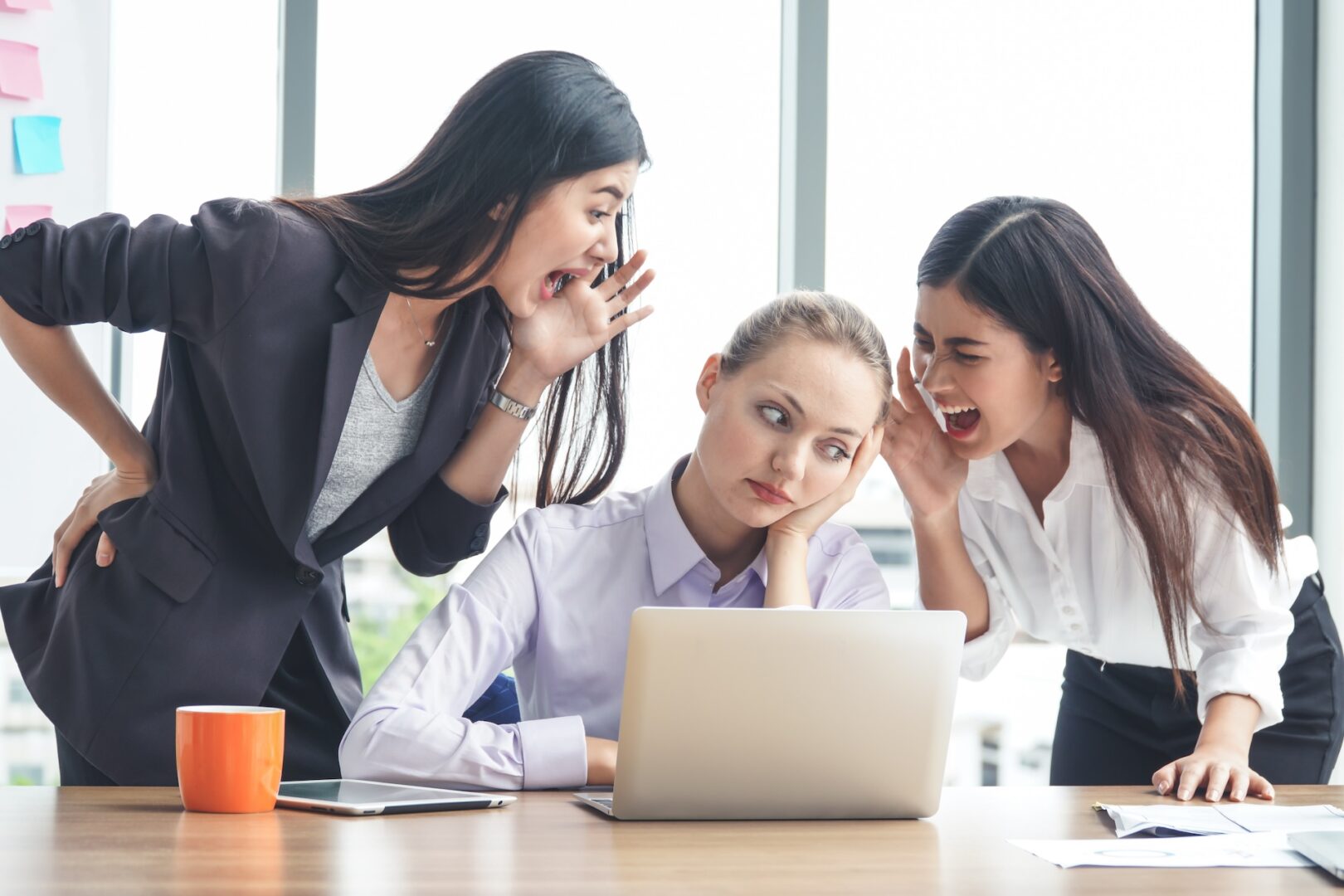 Three women are at a desk. The woman in the middle, looking bored, is using a laptop. The other two are on either side of her, leaning in and yelling. An orange coffee mug is on the desk, and large windows are in the background.