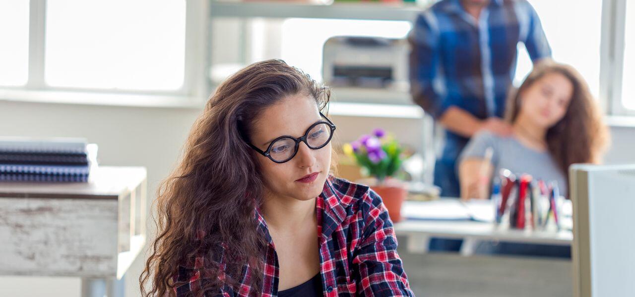 A woman with long curly hair and glasses sits at a desk, looking thoughtful. She wears a red plaid shirt. In the background, another woman and a man are visible, blurred, suggesting an office setting.