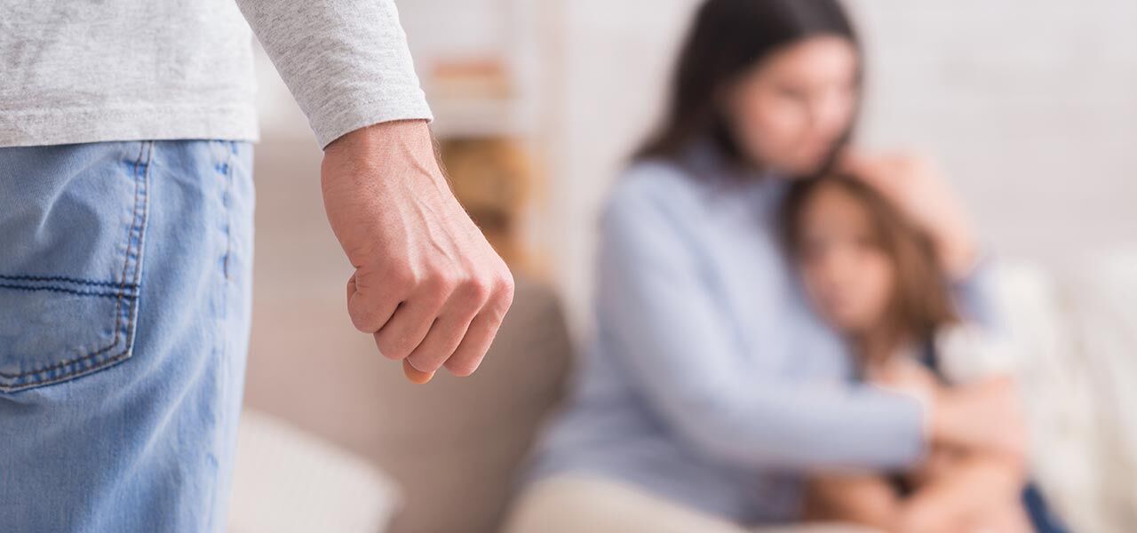 A woman and child sit on a couch, looking distressed. In the foreground, a person stands with a clenched fist, creating a tense atmosphere. The focus is on the woman's protective embrace of the child.