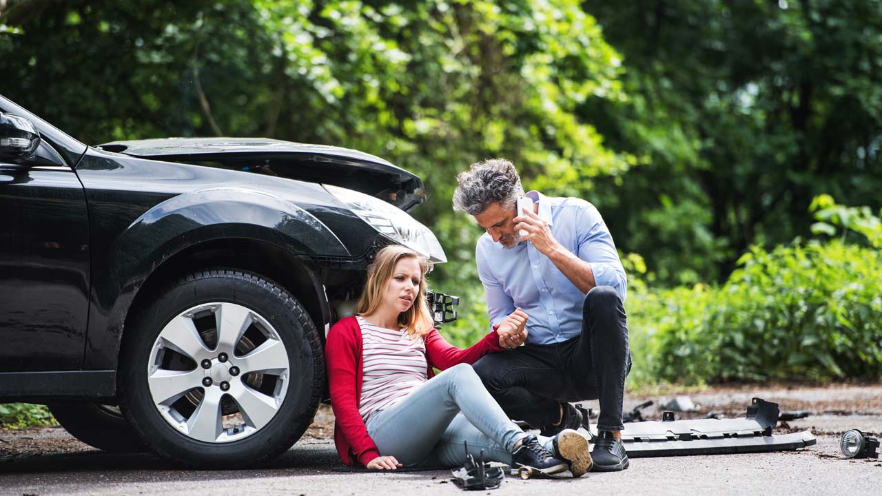 A car accident scene shows a man on the phone and a woman sitting on the ground next to a damaged black car. He appears to be assisting her, with scattered car parts visible nearby. Lush greenery surrounds the area.