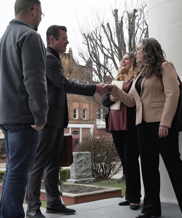 A group of four people stand outside a building. Two of them, a man in a dark suit and a woman in a light blazer, are shaking hands. Two others stand nearby, one man in a jacket and a woman with long hair, smiling. Trees and buildings are in the background.