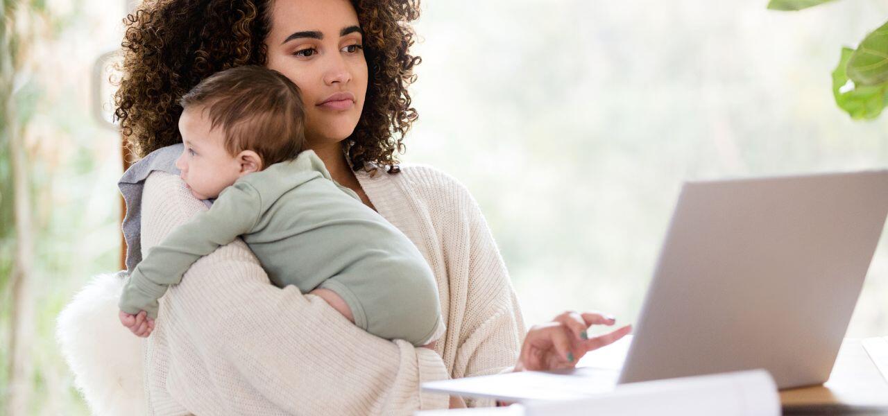 A woman with curly hair works on a laptop while holding a baby in her arms. She looks focused, and the baby is dressed in a green onesie. The setting is bright and airy, with a blurred background of greenery.