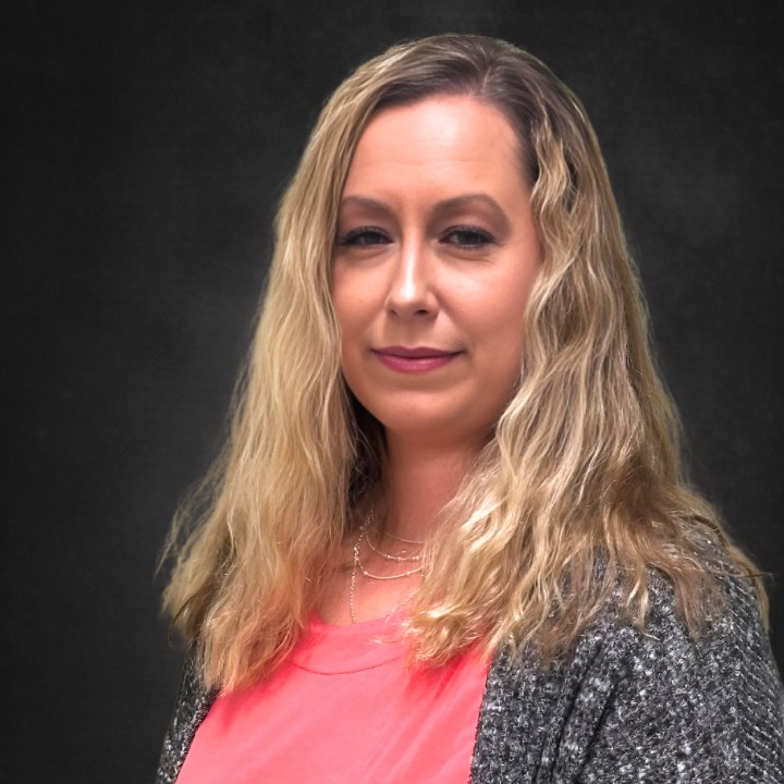 A woman with long, wavy blonde hair is standing against a dark background. She is wearing a pink top and a gray cardigan, looking at the camera with a neutral expression.