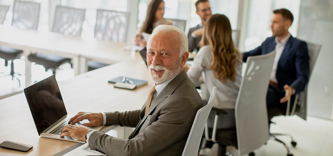 An older man in a suit smiles while typing on a laptop in a conference room. Four people are sitting and talking at the table in the background. The room is bright and modern.