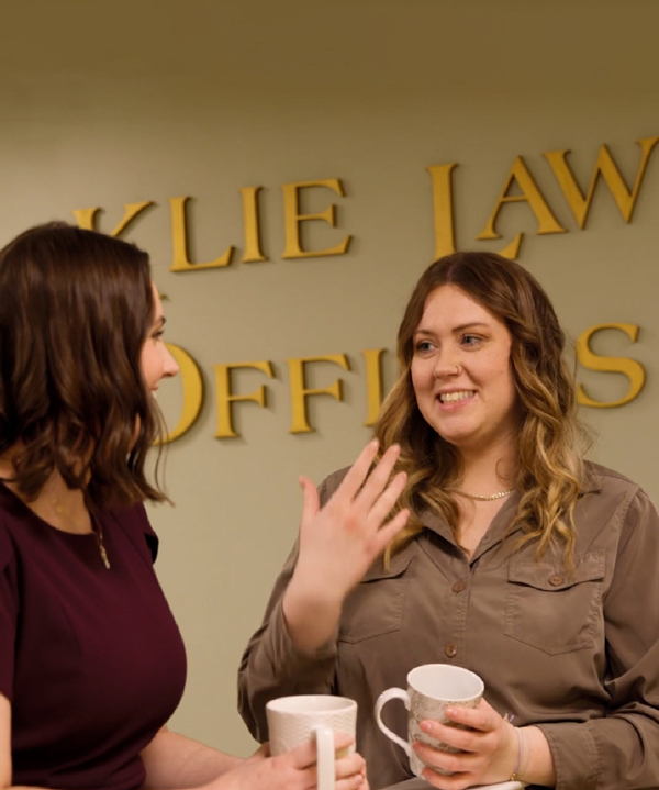 Two women are conversing and smiling while holding coffee mugs in front of a wall with partially visible text. The setting appears to be a professional office environment.