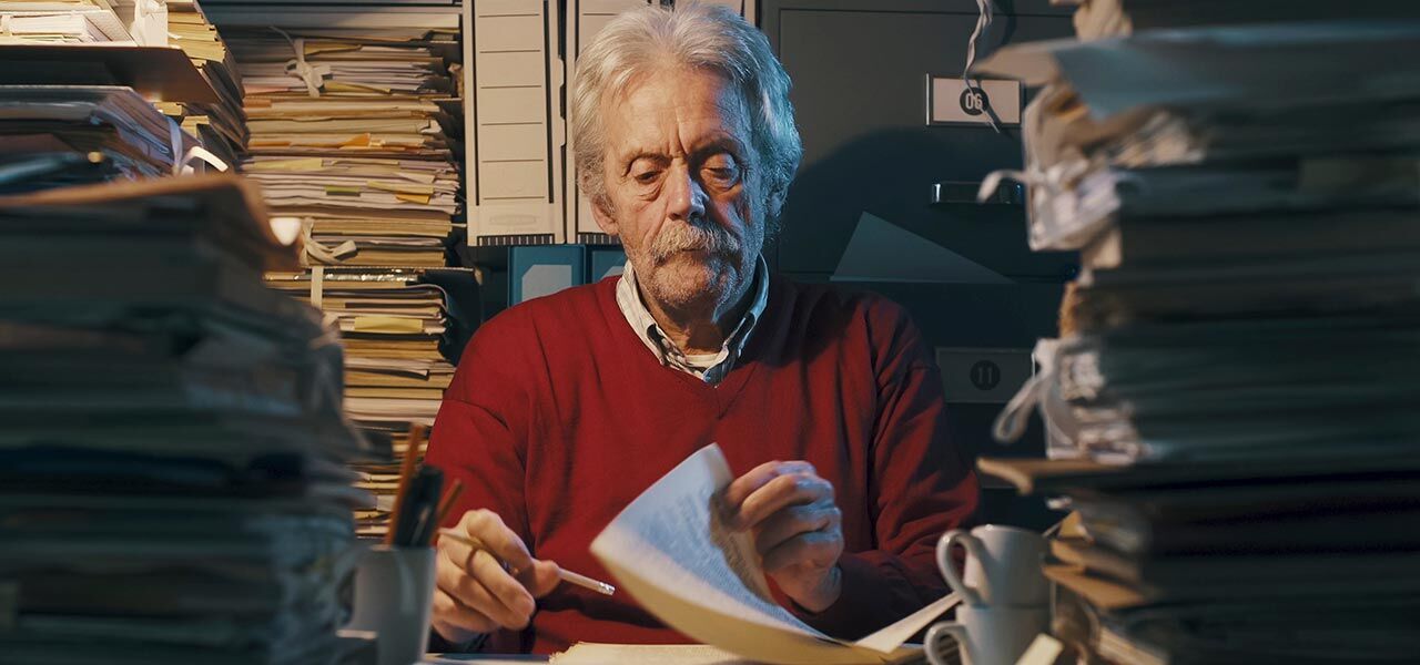 An elderly man with white hair and a mustache sits at a cluttered desk piled high with papers and documents. He wears a red sweater and is focused on reading a sheet of paper. The room is dimly lit.