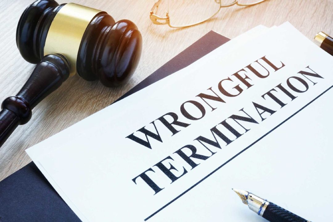 A wooden gavel rests beside a document labeled "Wrongful Termination" on a desk. A pair of glasses is partially visible in the background.