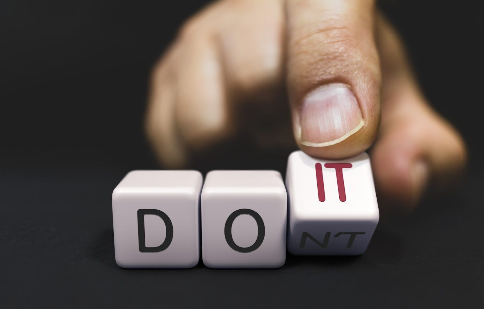 A hand is arranging lettered dice. The dice spell "DO" and "N'T," while the hand is changing "N'T" to "IT," creating the words "DO IT.
