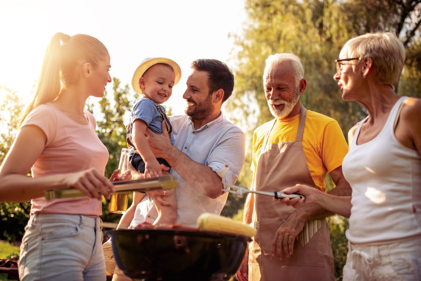 A family enjoys a sunny outdoor barbecue. A woman cooks at the grill while a man holds a smiling toddler wearing a hat. An elderly man in an apron and an older woman with glasses join in, standing together cheerfully.