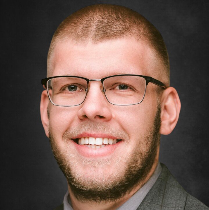 Portrait of a smiling man with a beard, short hair, and glasses, wearing a gray suit against a dark background.