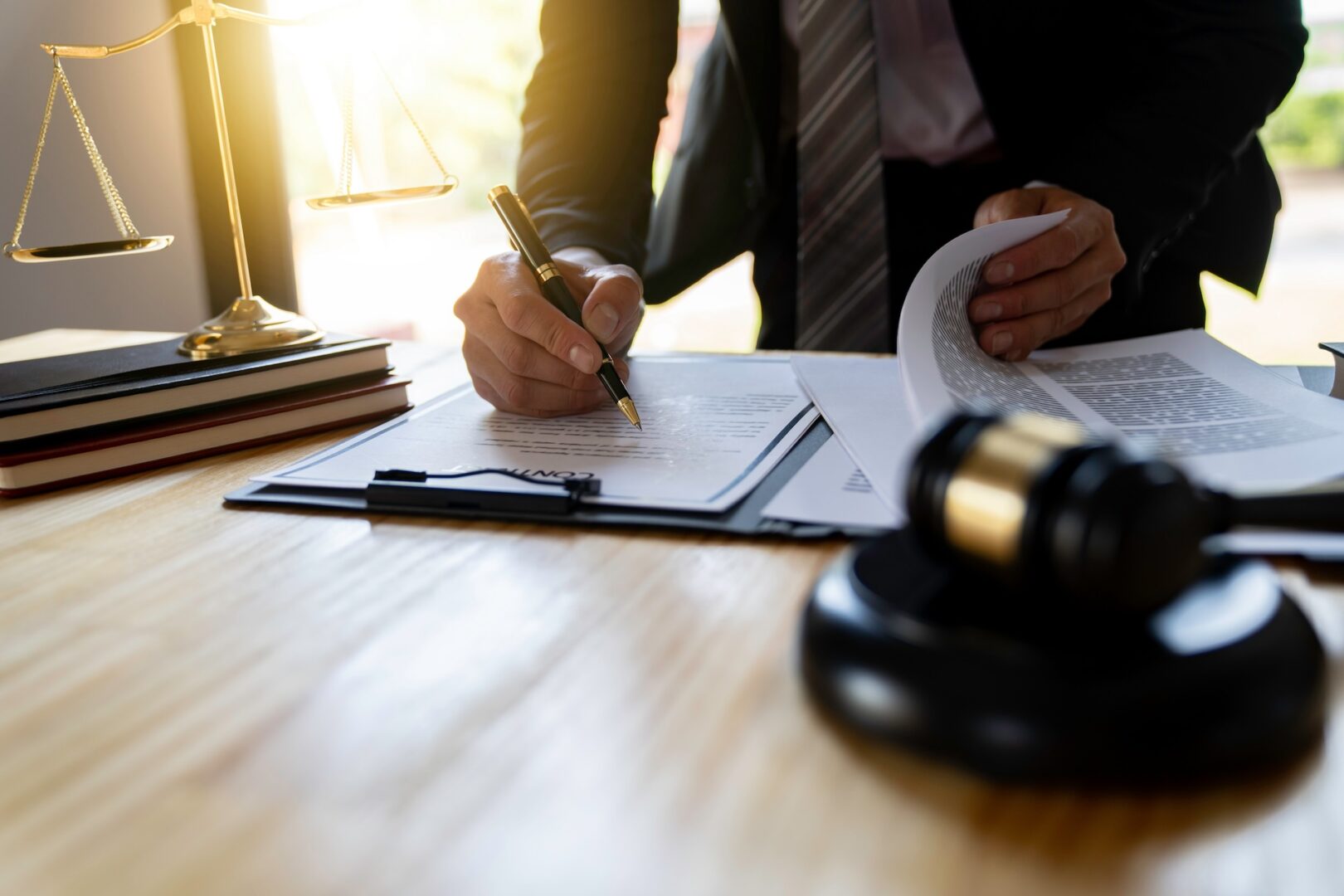 A person in a suit signs a document on a clipboard at a wooden desk, with books, scales of justice, and a gavel nearby. Sunlight illuminates the scene from the left side.