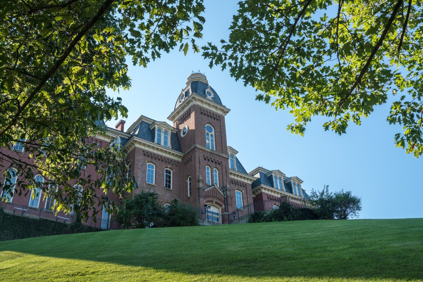 A historic brick building with a tall central tower and large windows sits atop a green hill. The sky is clear and blue, and tree branches frame the view from above.