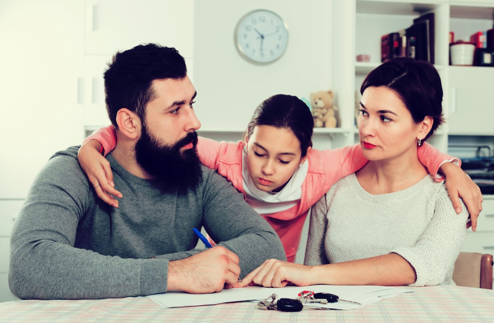 A serious-looking family sits at a table. The father is writing on a document, the mother listens intently, and the daughter wraps her arms around them, appearing thoughtful. A clock and shelves with items are in the background.