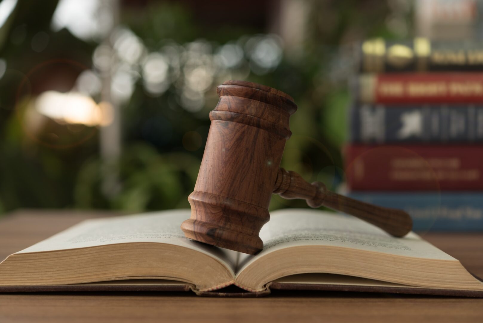 A wooden gavel rests on an open book, symbolizing justice and law. In the background, several stacked books are slightly blurred, along with greenery, creating a scholarly and natural ambiance.