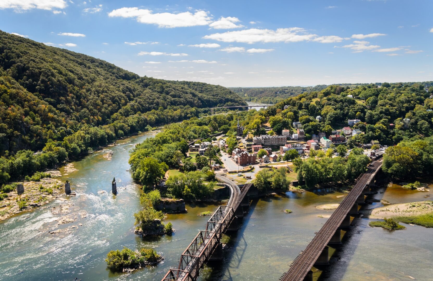 A scenic aerial view of a river curving through a lush, green valley. Bridges cross the water, connecting a small town nestled among trees. Rolling hills surround the area under a bright, sunny sky with scattered clouds.