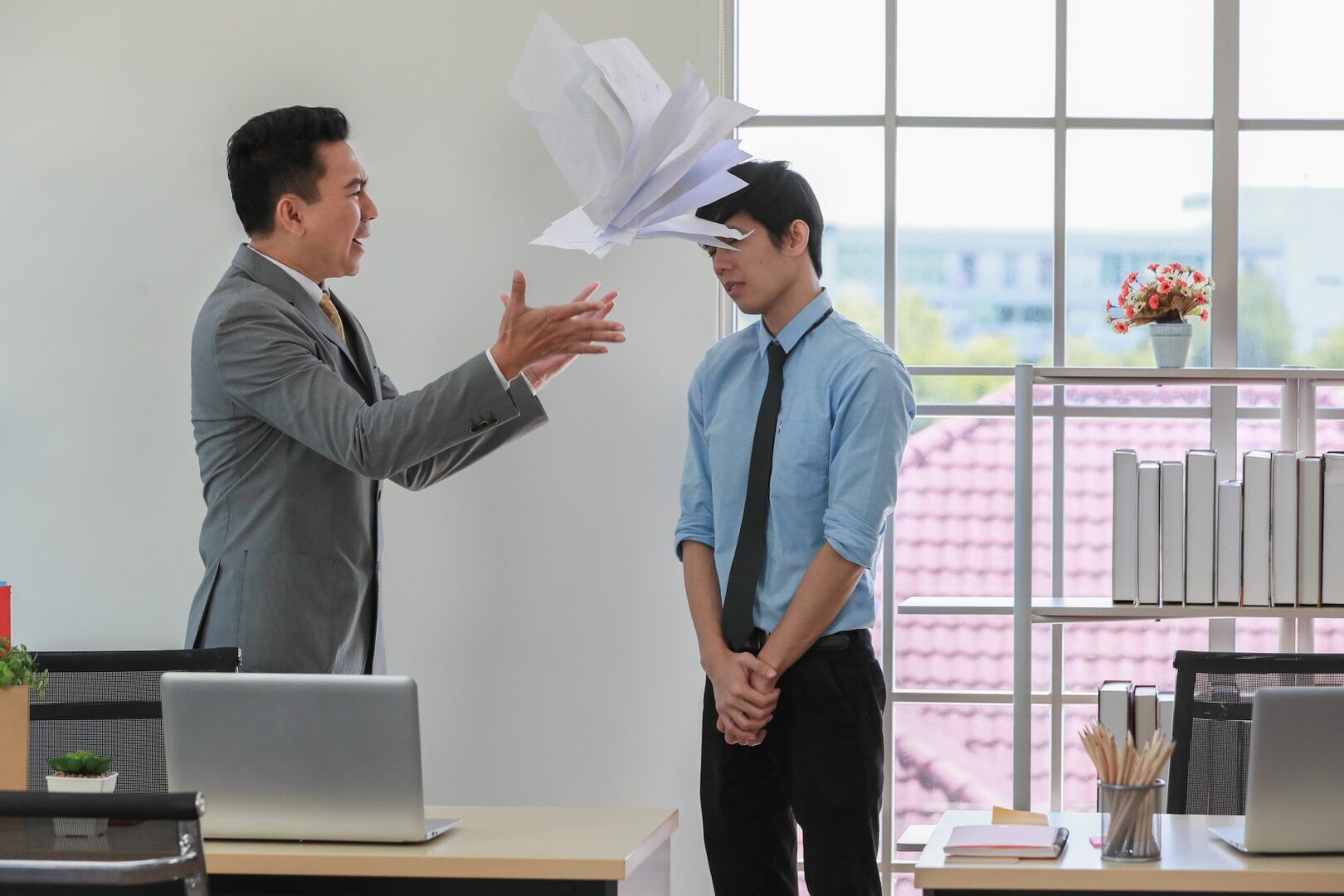 A man in a suit throws papers at another man in a blue shirt with a tie, who stands with his head down in an office. A laptop is on the desk, and a window with books and a potted plant is in the background.