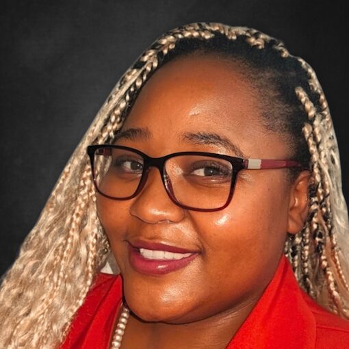 A woman with blonde braided hair and glasses smiles at the camera. She is wearing a red top and has a pearl necklace. The background is a dark, neutral color.