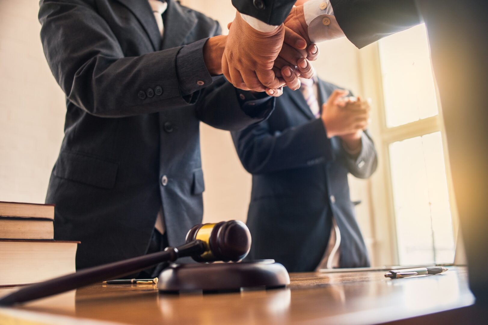 Two people in business suits shaking hands over a desk with a gavel in focus. Books and a pen are on the desk. Another person in the background is clapping. Sunlight streams through a window, creating a bright atmosphere.