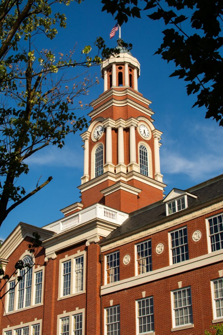 A red-brick building with a clock tower topped by a cupola and a small American flag. The sky is clear and blue, and tree branches frame the structure, adding a natural element to the architectural scene.