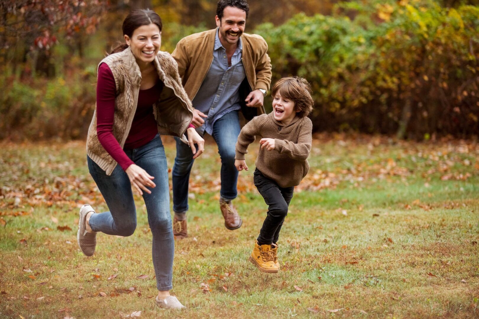 A family of three joyfully running through a grassy area during autumn. A woman and man chase a smiling young boy, surrounded by colorful fallen leaves and trees in the background.