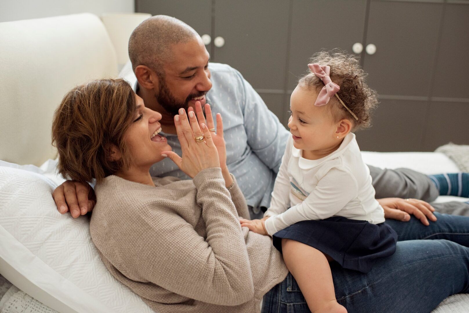 A joyful family scene: a woman and a man are lying on a bed, playfully interacting with a small child sitting on the woman's lap. They're smiling, and the child has a pink bow in her curly hair. The room is cozy with a soft background.