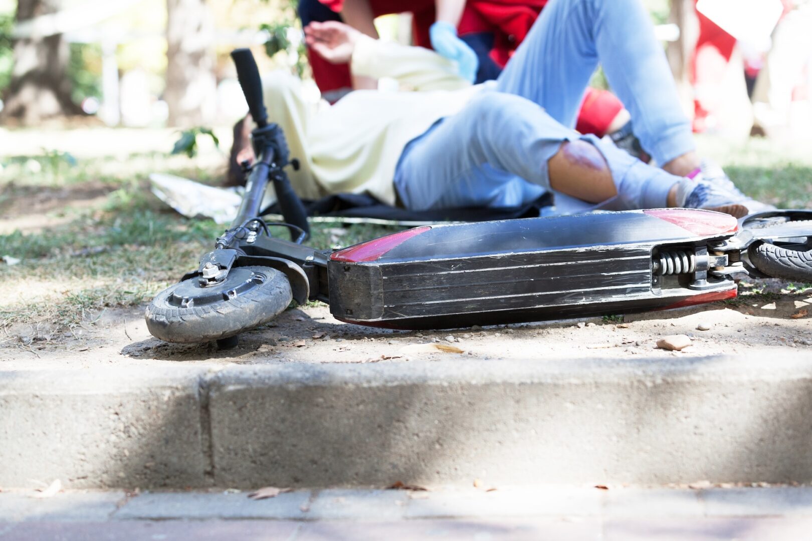 A person lying on the ground beside a fallen electric scooter on a sidewalk. Their jeans are ripped at the knee, showing a bandaged wound. The scene is in a park setting with green grass and a blurred background.