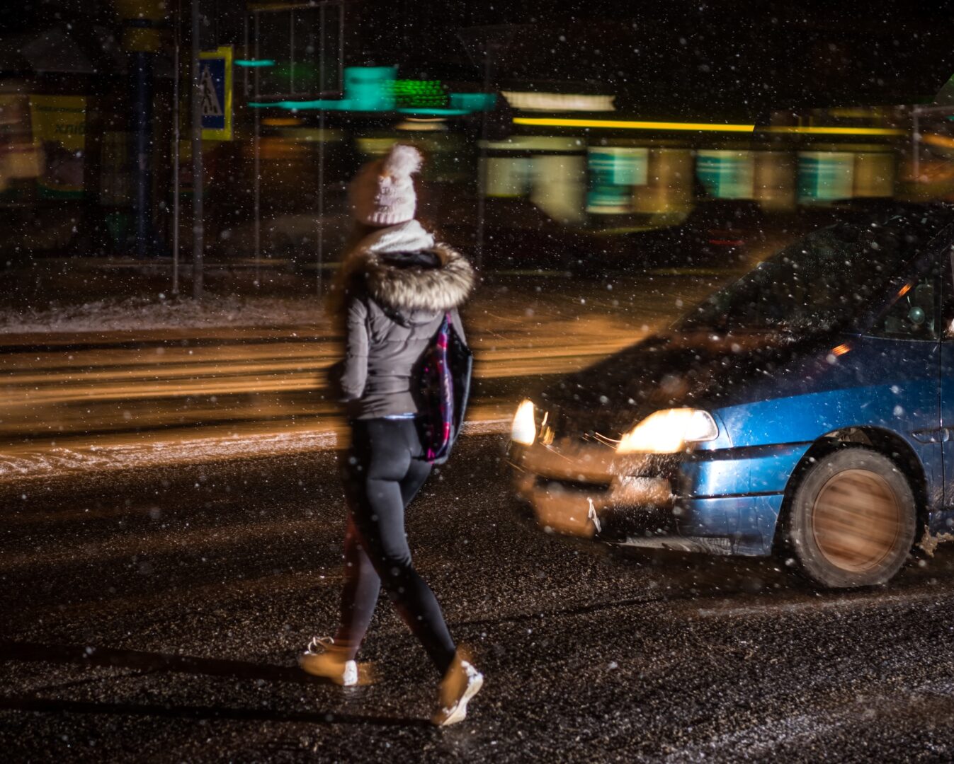 A person in winter attire crosses a snowy street at night, illuminated by streetlights. A blue car drives past, with snowflakes visible against the dark background, creating a scene of motion and contrast.