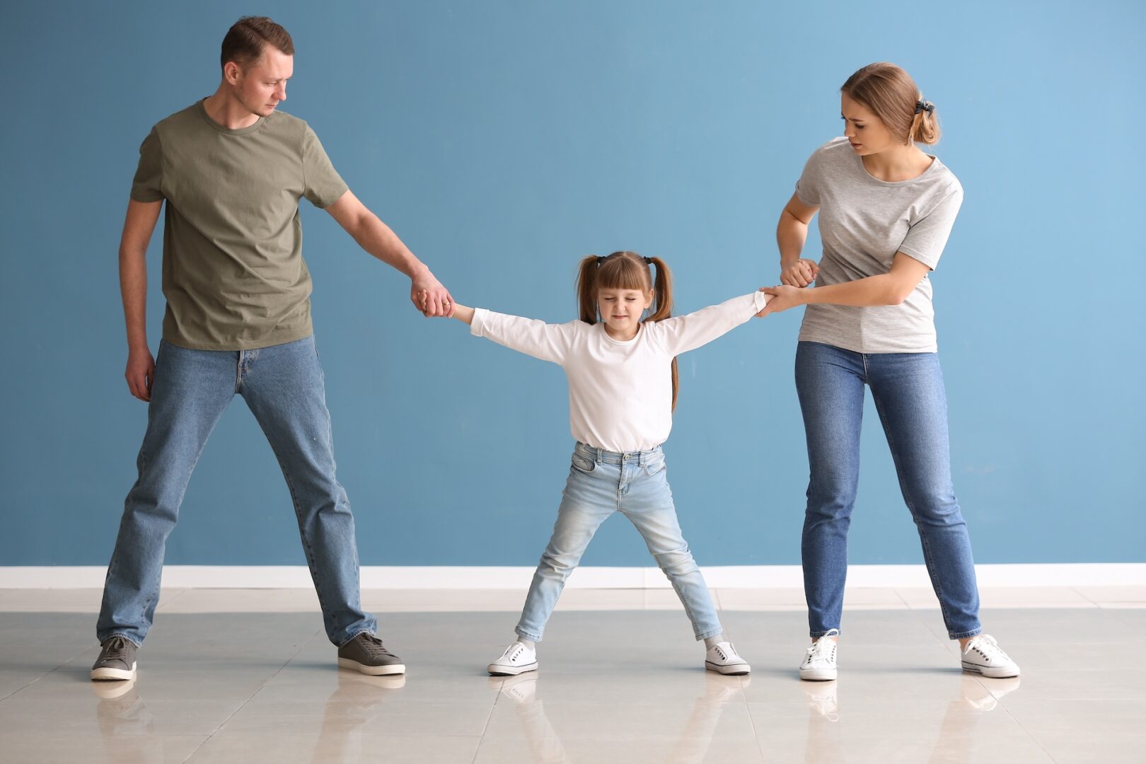 A young girl with pigtails playfully stretches her arms between an adult man and woman holding her hands. The man in a green shirt and the woman in a gray shirt stand on either side, smiling against a blue background.