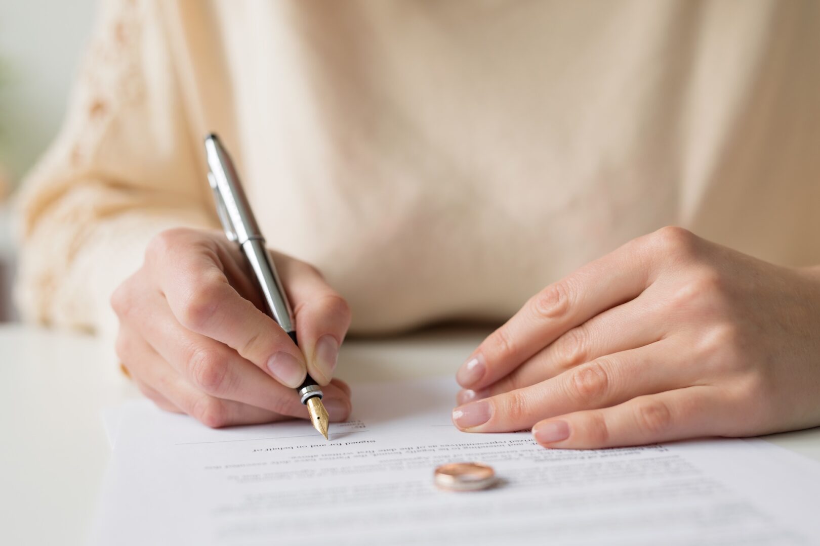 A person writing on a document with a silver pen. A pair of wedding rings rests on the table next to the paper. The person wears a light beige shirt, and their nails are neatly manicured. The scene suggests a signing of an important agreement.