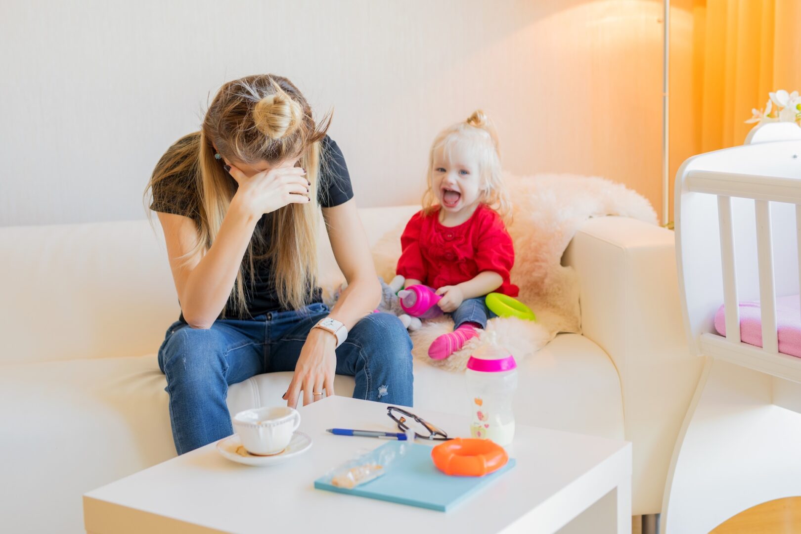 A woman sits on a couch with her head in her hand, looking stressed. Beside her, a smiling child with a toy sits happily. A table in the foreground holds a cup, a book, and other items. The room is well-lit and cozy.