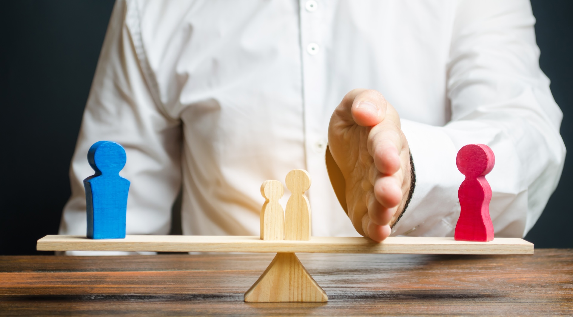 A person in a white shirt places a hand vertically between blue and red wooden figurines on a wooden seesaw. Two smaller figures stand in the center. The setup symbolizes conflict resolution or mediation.