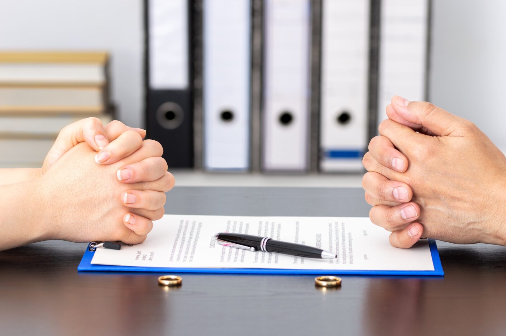 Two pairs of hands rest on a table, clasped near a document with a pen on top. A gold ring lies on each side of the paper. In the background, there are stacked folders and binders.