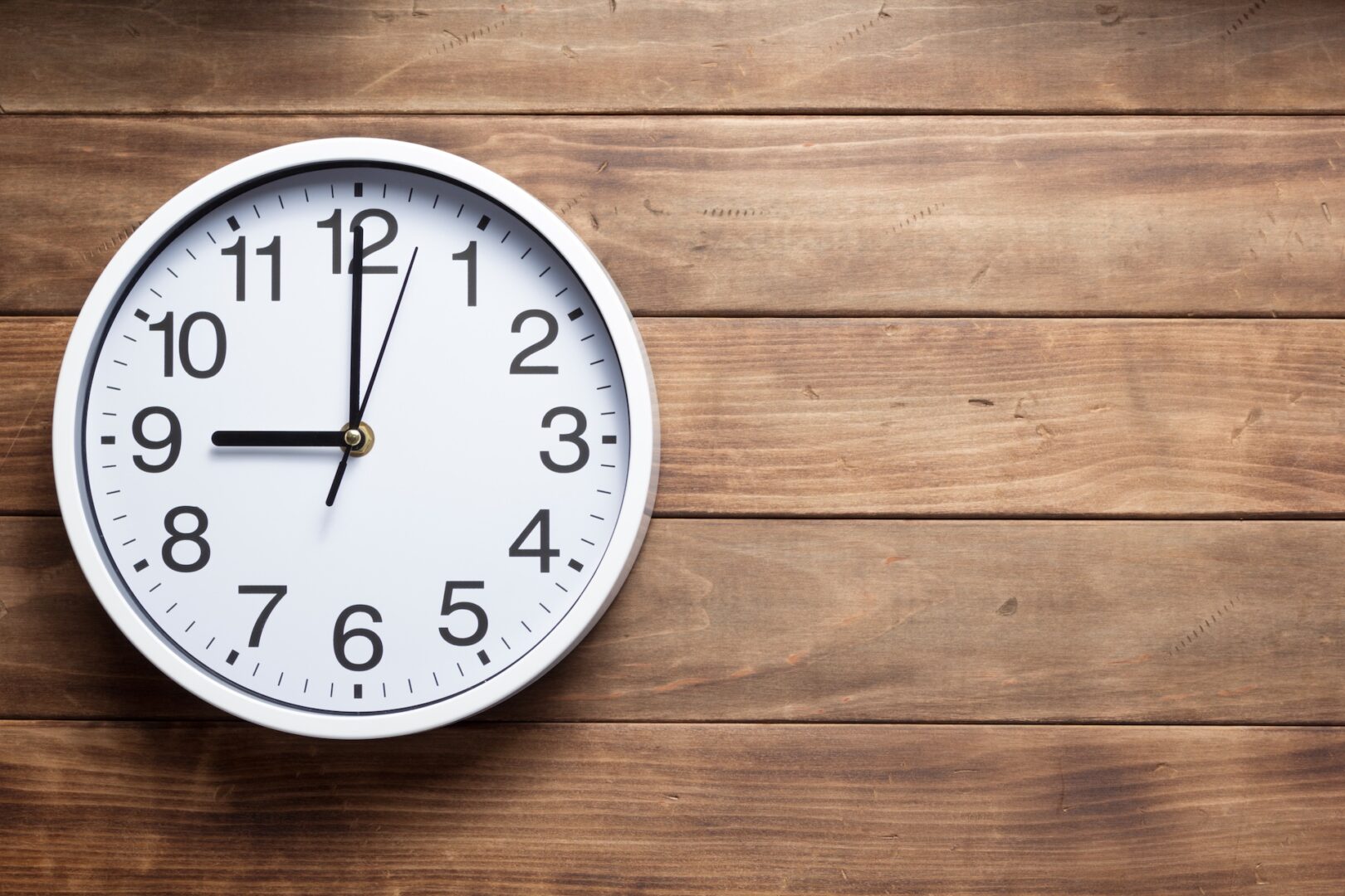 A round wall clock with a white face and black numbers and hands displays 10:09. It is mounted on a wooden plank wall.