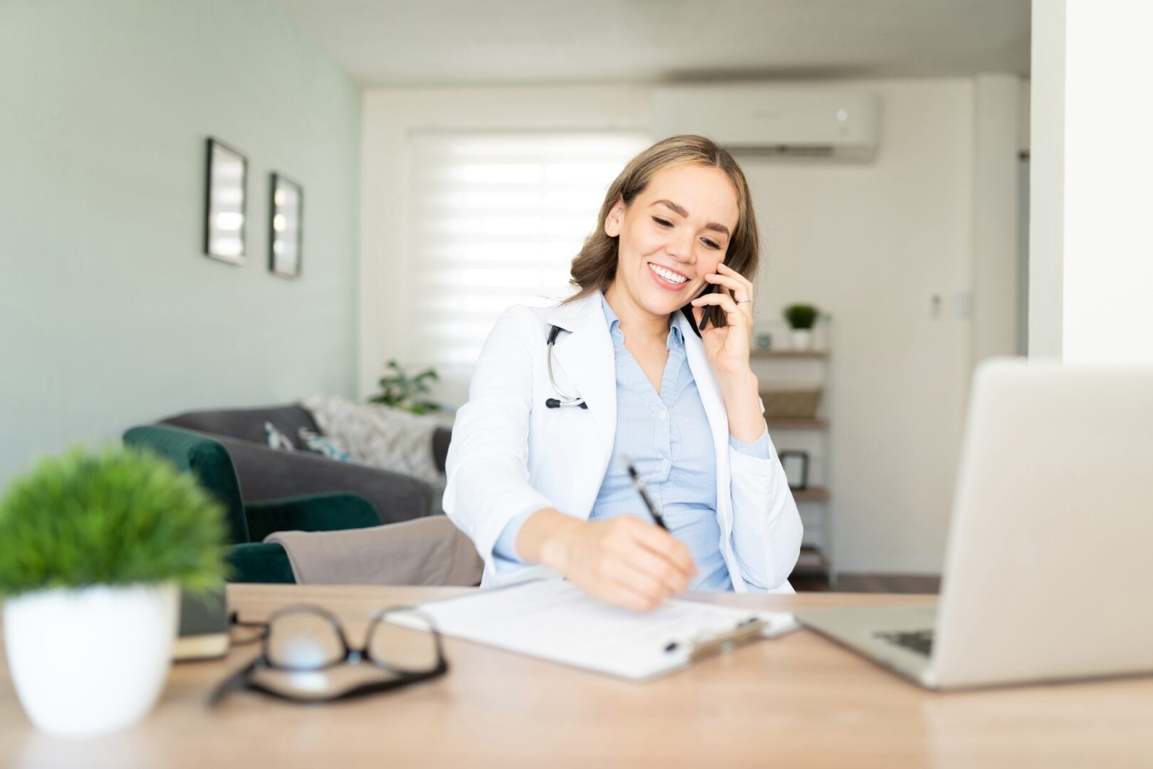 A smiling woman in a white coat sits at a desk, talking on the phone and writing on a clipboard. A laptop, glasses, and a small plant are in front of her. The room has a couch and framed pictures on the wall.