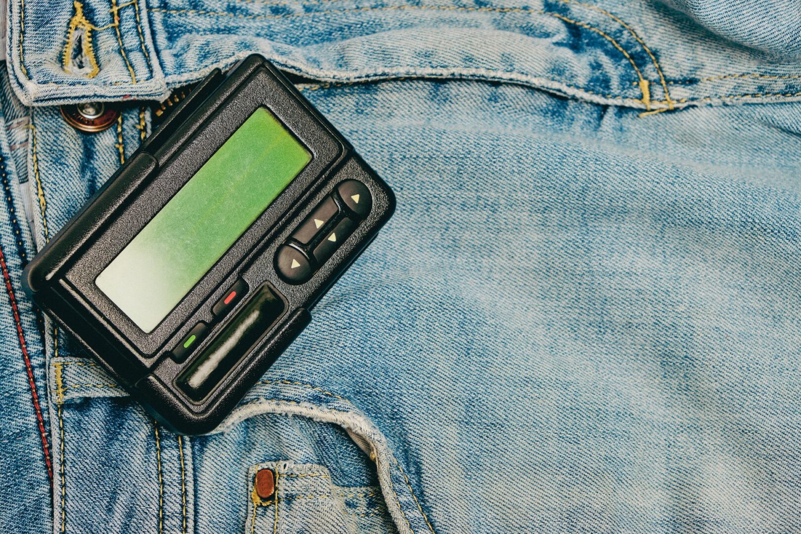 A vintage black pager with a green screen and buttons lies on faded blue denim fabric, positioned near a jean pocket. The fabric has visible seams and a button.