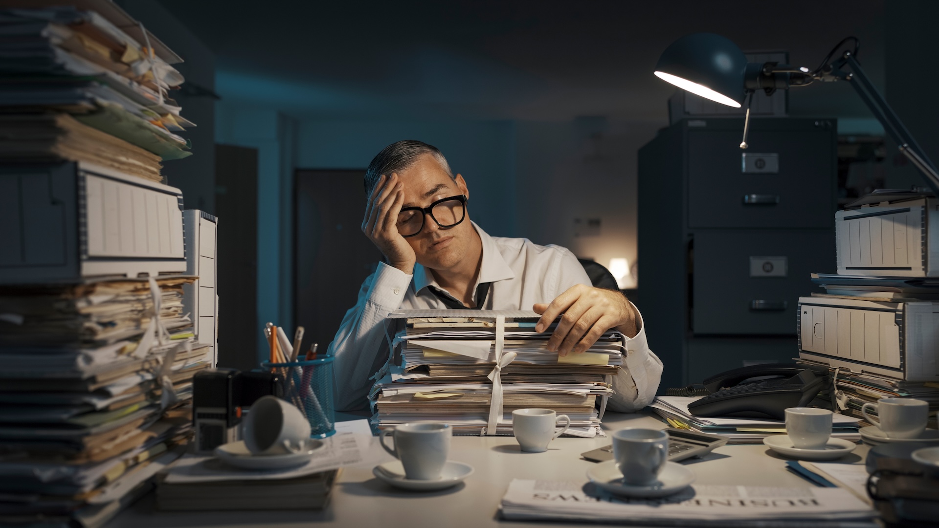 A man with glasses and a tired expression sits at a cluttered desk, resting his head on his hand. The desk is filled with stacks of paperwork, coffee cups, and a desk lamp. The scene is dimly lit, suggesting evening or night-time work.