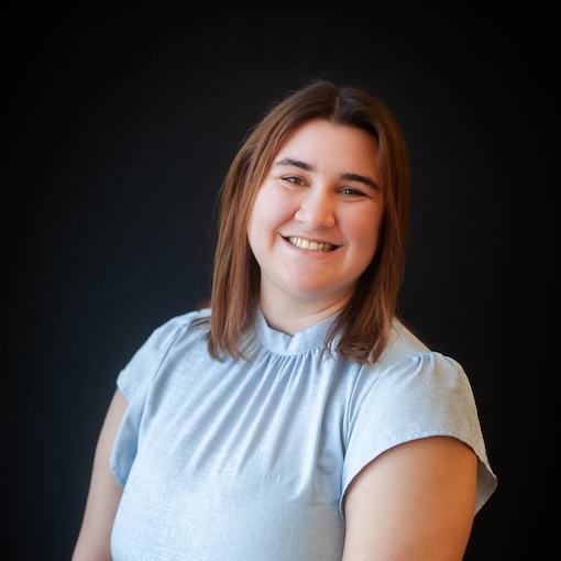 Smiling person with shoulder-length brown hair wearing a light blue top against a dark background.