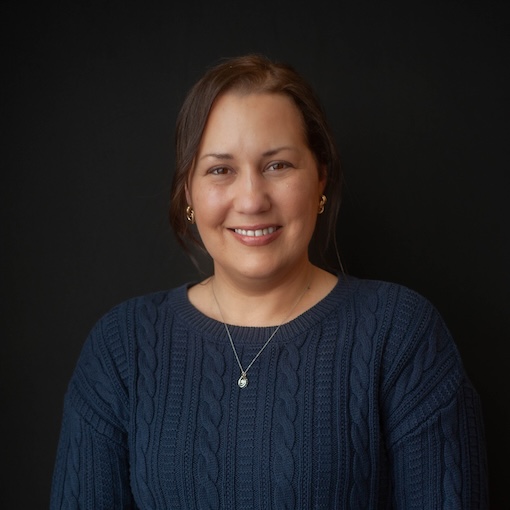 A person with long hair is smiling, wearing a navy blue sweater and a necklace. The background is dark.