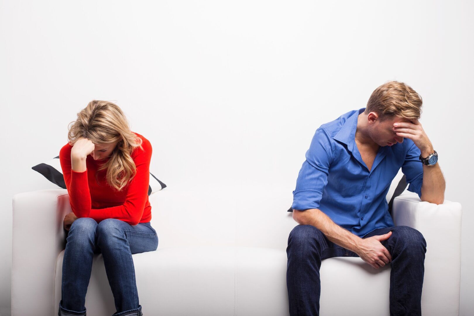 A woman and a man sit apart on a white couch, both with heads down and hands on their foreheads, appearing upset or stressed—perhaps reflecting on what is the difference between legal separation and divorce. The background is plain white.