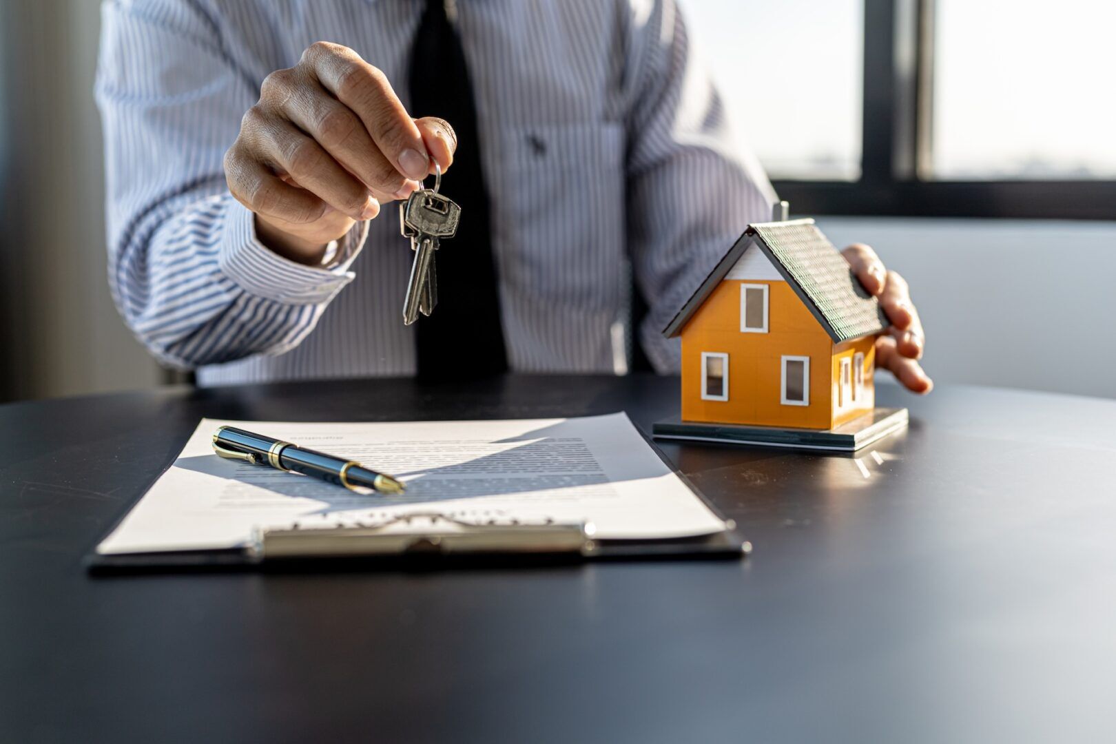 A person in business attire holds a set of keys over a desk with a contract, pen, and small model house, symbolizing real estate or home purchase—key elements when considering what property can be divided in divorce.