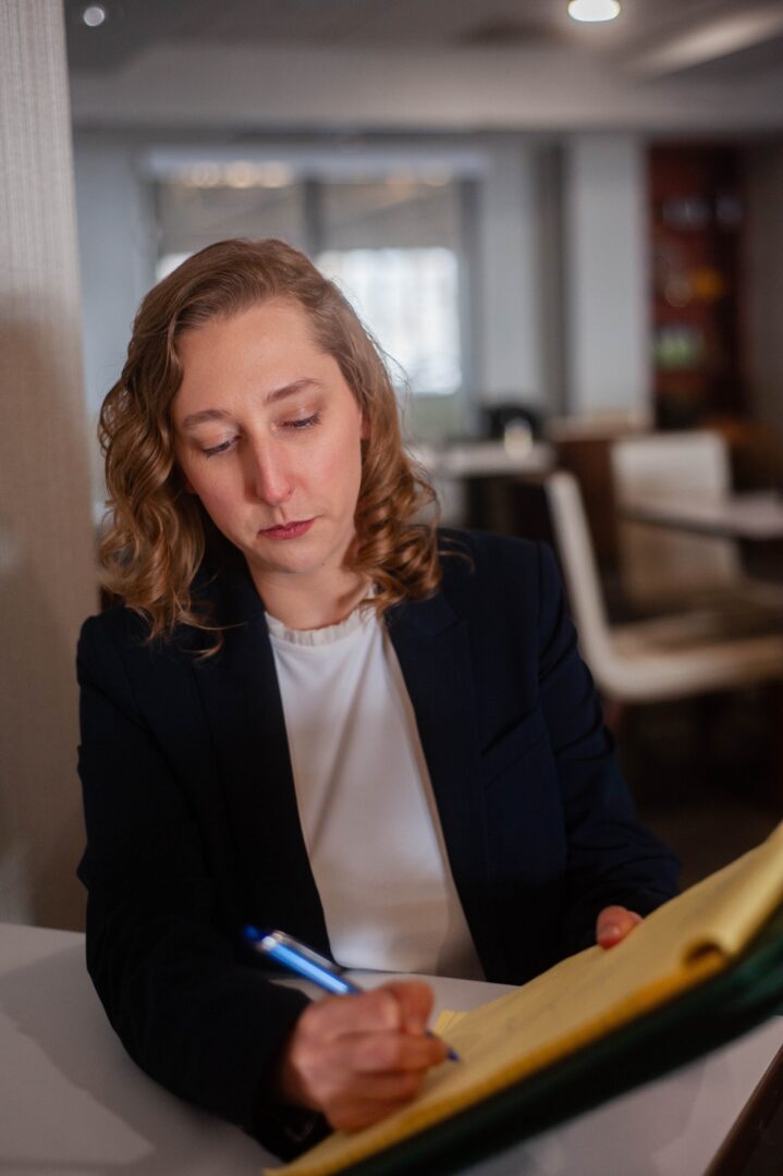 A person with wavy, shoulder-length hair wearing a dark blazer and white shirt sits at a table, writing notes on a yellow legal pad with a blue pen in a well-lit indoor setting. A person with wavy, shoulder-length hair wearing a dark blazer and white shirt sits at a table, writing notes on a yellow legal pad with a blue pen in a well-lit indoor setting.