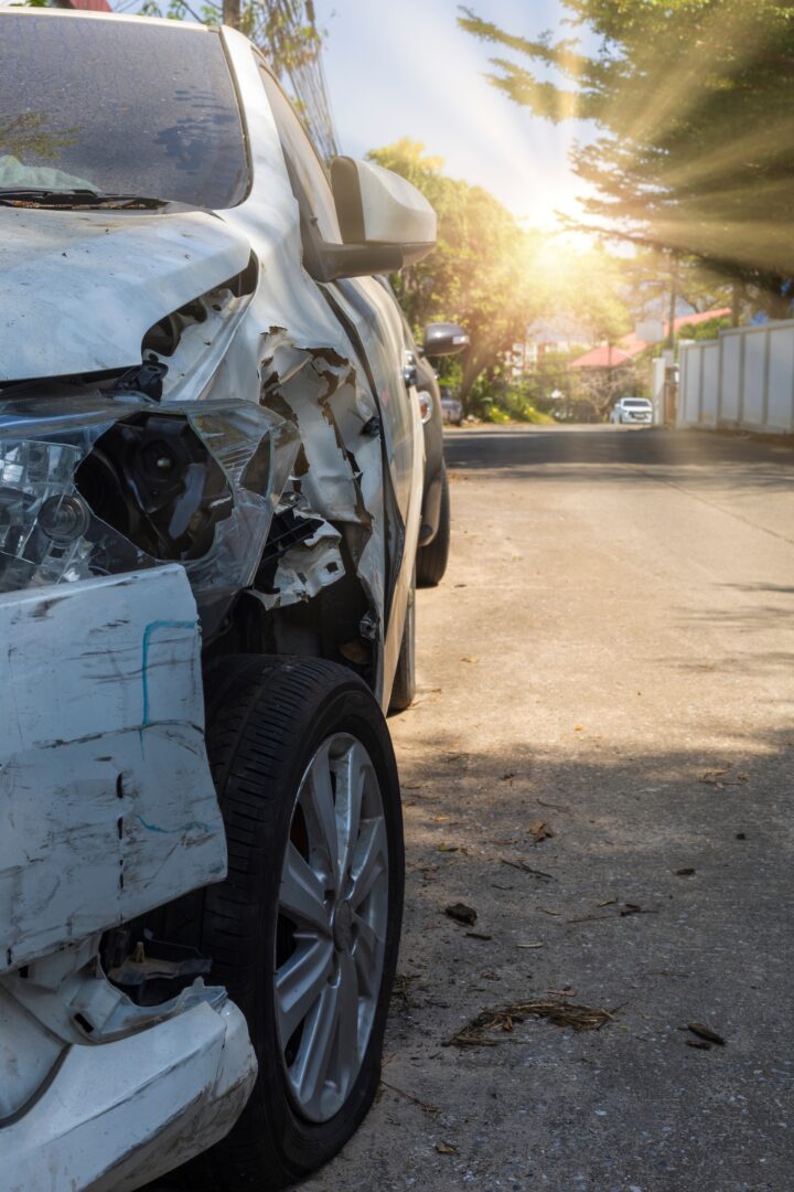 A white car with significant front-end damage is parked on the side of a sunlit street. The headlight and fender are smashed, and the bright sun shines through trees in the background. A white car with significant front-end damage is parked on the side of a sunlit street. The headlight and fender are smashed, and the bright sun shines through trees in the background.