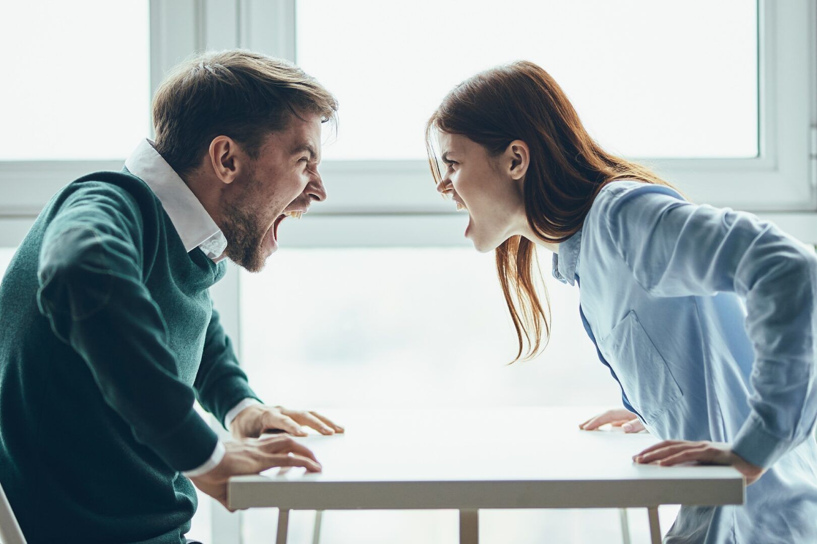 A man and a woman sit across from each other at a table, leaning forward and yelling with angry expressions—a tense scene that raises the question: What Is Necessary to File a Temporary Restraining Order in West Virginia? The background is bright with large windows.