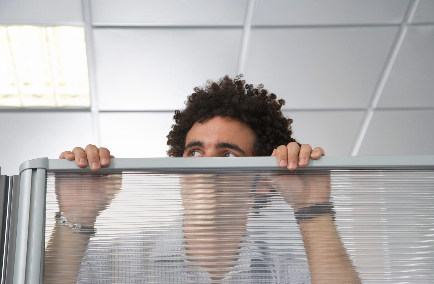 A person with curly hair peers nervously over a frosted glass office divider, gripping the top edge with both hands—perhaps anxious about workplace discrimination in West Virginia. The ceiling and fluorescent lights are visible above.