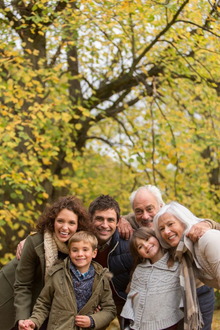 Three generations of a family—two children, their parents, and grandparents—smiling and posing closely together outdoors in a park with autumn leaves in the background. Three generations of a family—two children, their parents, and grandparents—smiling and posing closely together outdoors in a park with autumn leaves in the background.