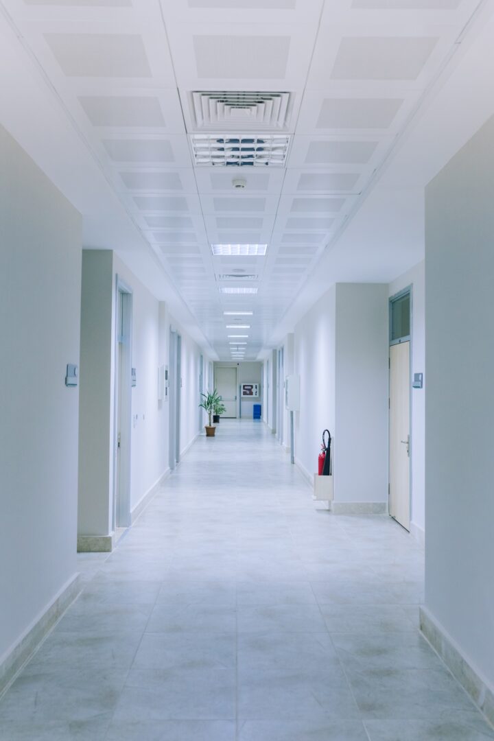A long, empty hallway in a modern building with white walls, tiled floor, doors on both sides, a ceiling with vents and lights, a fire extinguisher, and a potted plant visible at the far end. A long, empty hallway in a modern building with white walls, tiled floor, doors on both sides, a ceiling with vents and lights, a fire extinguisher, and a potted plant visible at the far end.
