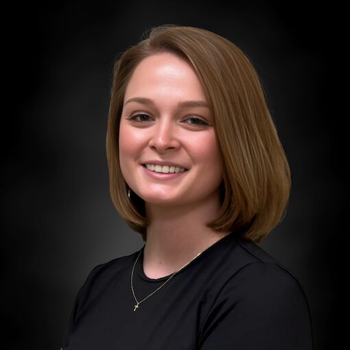 A woman with straight, shoulder-length light brown hair smiles at the camera. She is wearing a black top and a delicate necklace with a small pendant, set against a dark, blurred background.