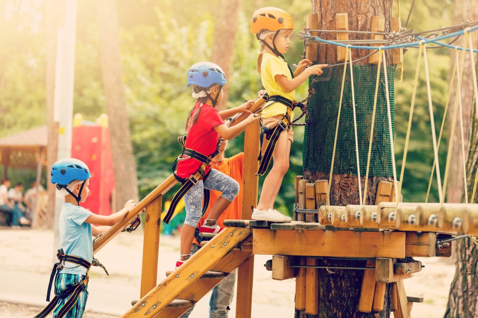 Three children wearing helmets and harnesses climb a wooden structure and prepare to cross a rope bridge in an outdoor adventure park surrounded by trees, reminding visitors to stay safe—know what to do if hurt in a slip and fall accident.