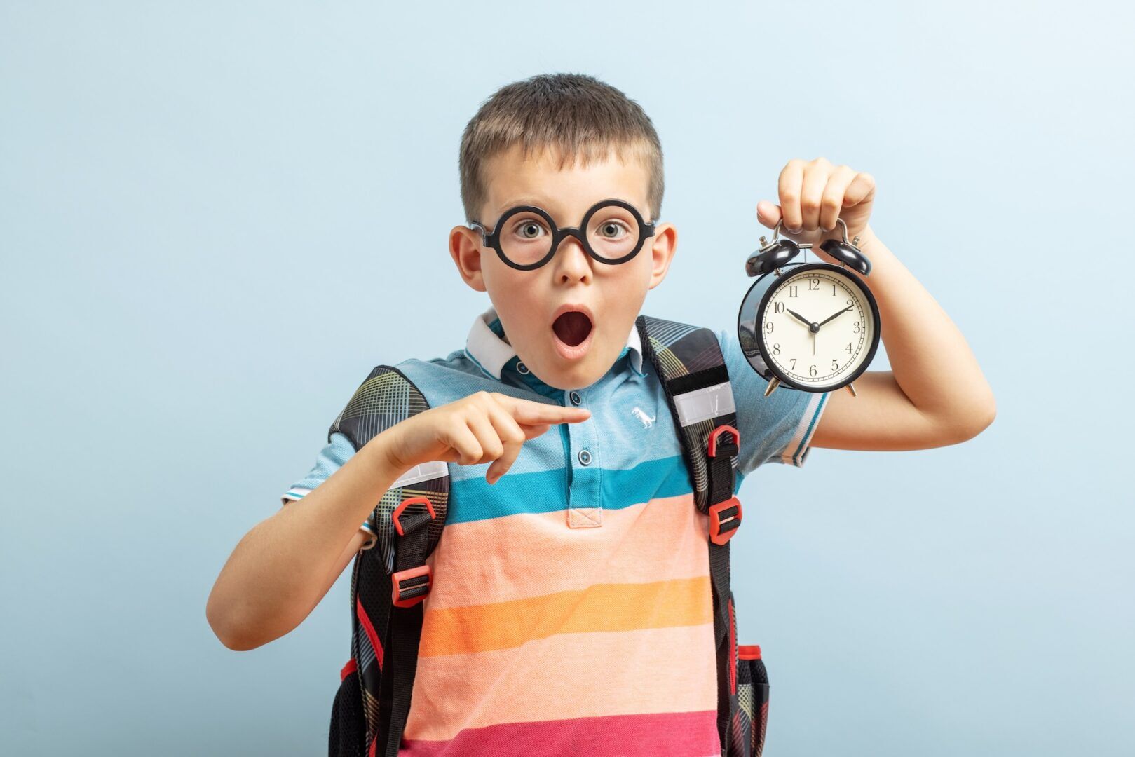 A surprised boy wearing glasses and a colorful striped shirt points at an alarm clock he is holding, with a blue background. He has a backpack on and looks amazed by the time—just like learning about Understanding Child Custody Rights in WV.